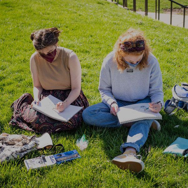 Two students sit outside drawing for their costume design class.